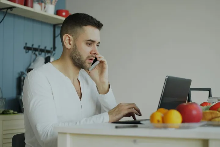 Man at his kitchen table on the phone with a business, his laptop open in front of him