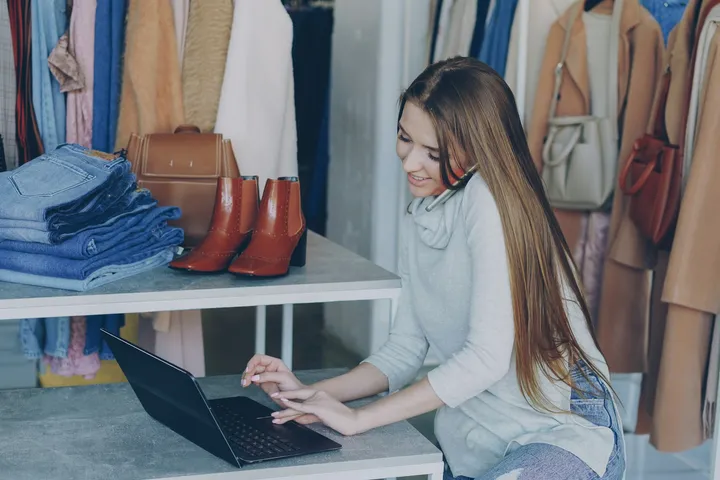Clothing store owner working on her product catalog on a laptop between racks of coats and shelves of jeans