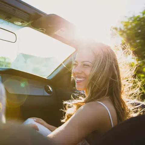 A smiling driver in the sun behind a clear windshield, the trees outside sharp through the glass