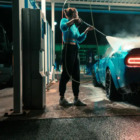 A woman spraying her blue car with a pressure wand at a self-serve car wash bay at night