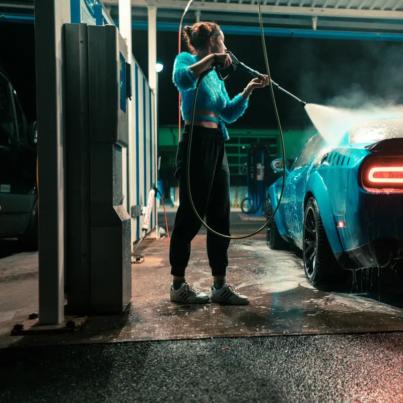 Email header photo: a woman spraying her blue car with a pressure wand at a self-serve car wash bay at night