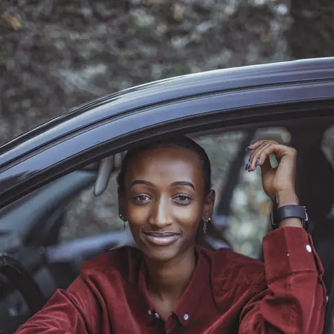 A smiling woman in a red corduroy shirt leaning on the open window of a dark car