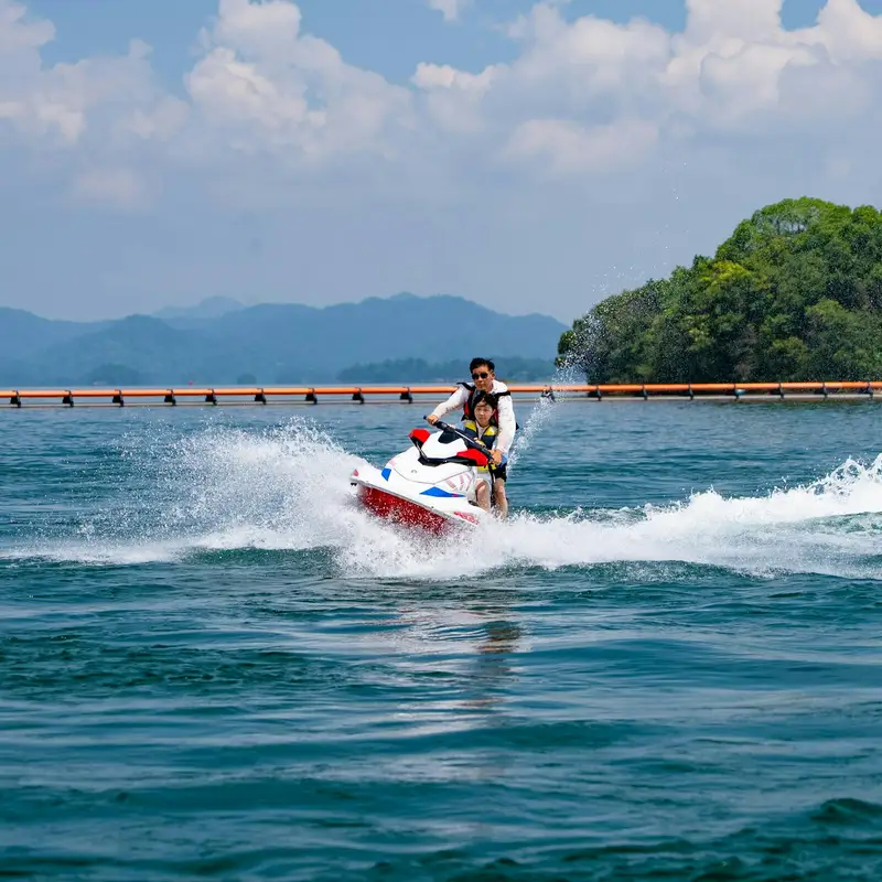 Email header photo: two riders in life jackets on a red and white personal watercraft throwing spray across a lake, with a wooded island and mountains behind