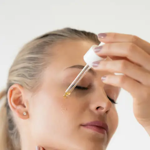 A woman with her eyes closed applies a drop of facial serum to her forehead from a glass dropper