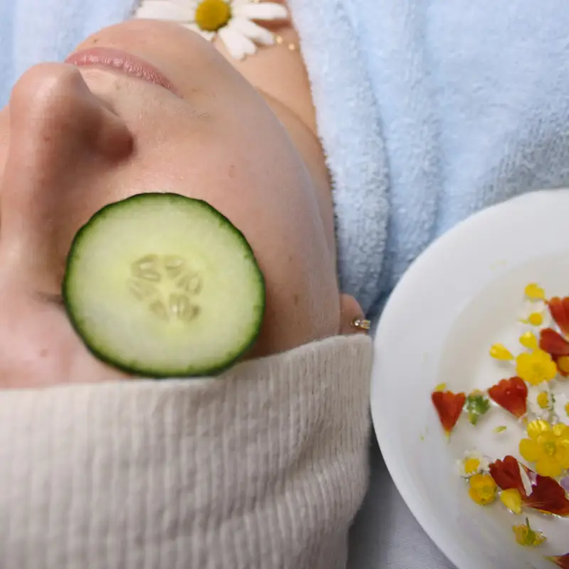 Email header photo: spa guest in a soft blue robe resting with a cucumber slice over one eye and a daisy beside her face