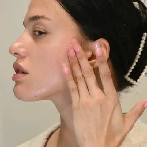 Young woman with dark hair pinned back touching her dewy, freshly cared-for cheek with her fingertips