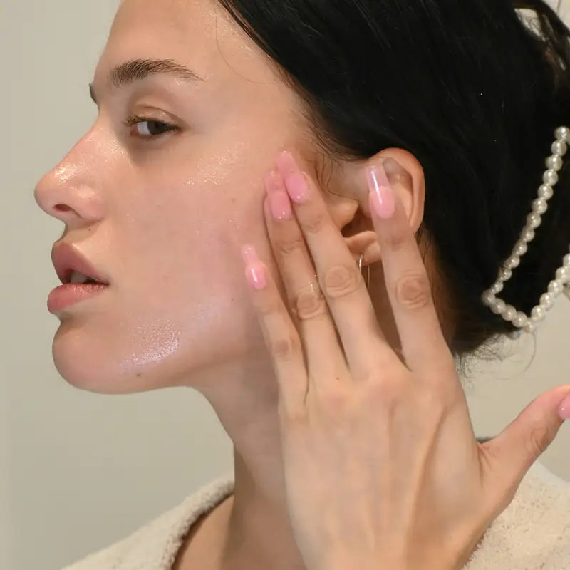 Email header photo: young woman with dark hair pinned back touching her dewy, freshly cared-for cheek with her fingertips