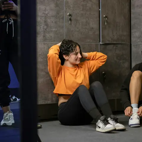 Two women sitting on a locker room floor after a workout, one tying her shoelaces