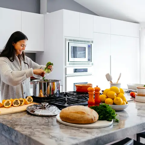 Smiling woman adding fresh greens to a pot in a bright home kitchen, with bread, lemons and vegetables on the counter