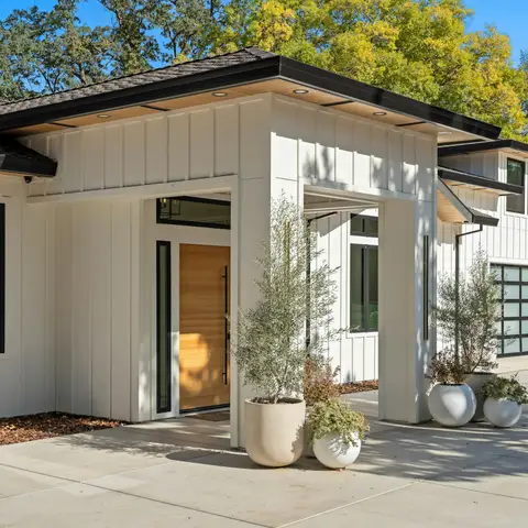 A freshly finished white board-and-batten home with a wood front door, potted olive trees and a clean concrete drive