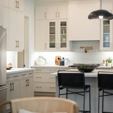 A spotless white kitchen with a clear island, black bar stools and a brass faucet under soft pendant light