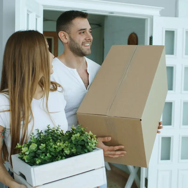 Email header photo: a smiling couple carrying a moving box and a crate of plants through the door of their new home