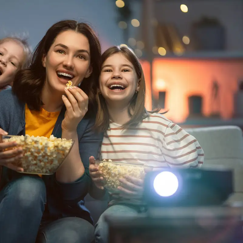 Email header photo: a mother and two daughters laughing on a sofa with bowls of popcorn during a movie night, lit by a projector in a dim room