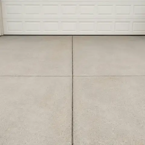 A clean, light gray concrete driveway with neat control joints leading to a white garage door set in a brick wall