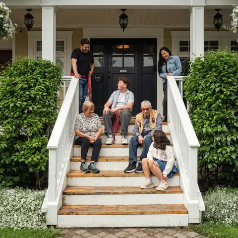 Three generations of a family sitting and standing on the white front porch steps of a home, framed by hanging flower baskets