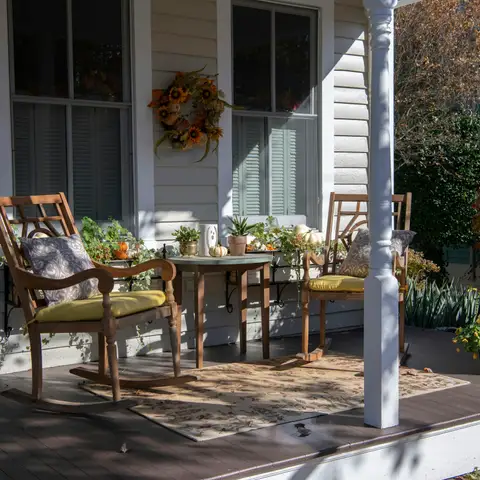 A covered front porch with white columns, two wooden chairs, a small table, potted plants and an autumn wreath on the window