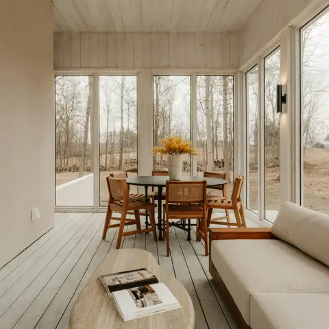 A finished four-season room with a fireplace, a cream sofa, a wooden dining table and floor-to-ceiling windows onto bare winter trees