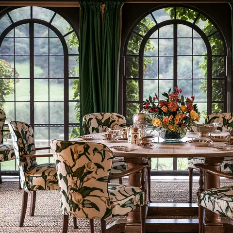 A designed dining room with floral upholstered chairs around a round wood table, green drapes and tall arched windows