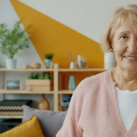 Older woman with short blond hair in a pink cardigan smiling in a bright living room