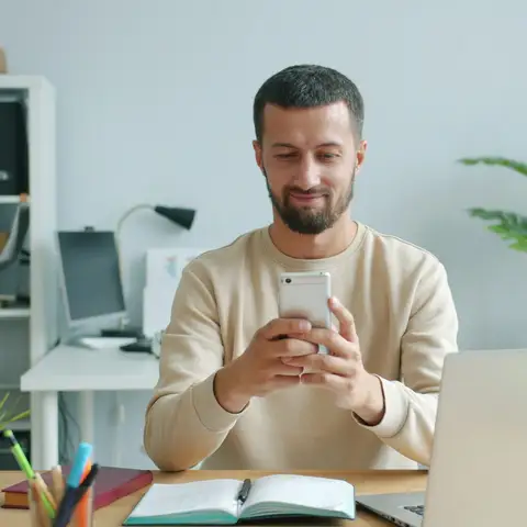Bearded man in a cream sweater smiling at his phone at a desk with a laptop and potted plants