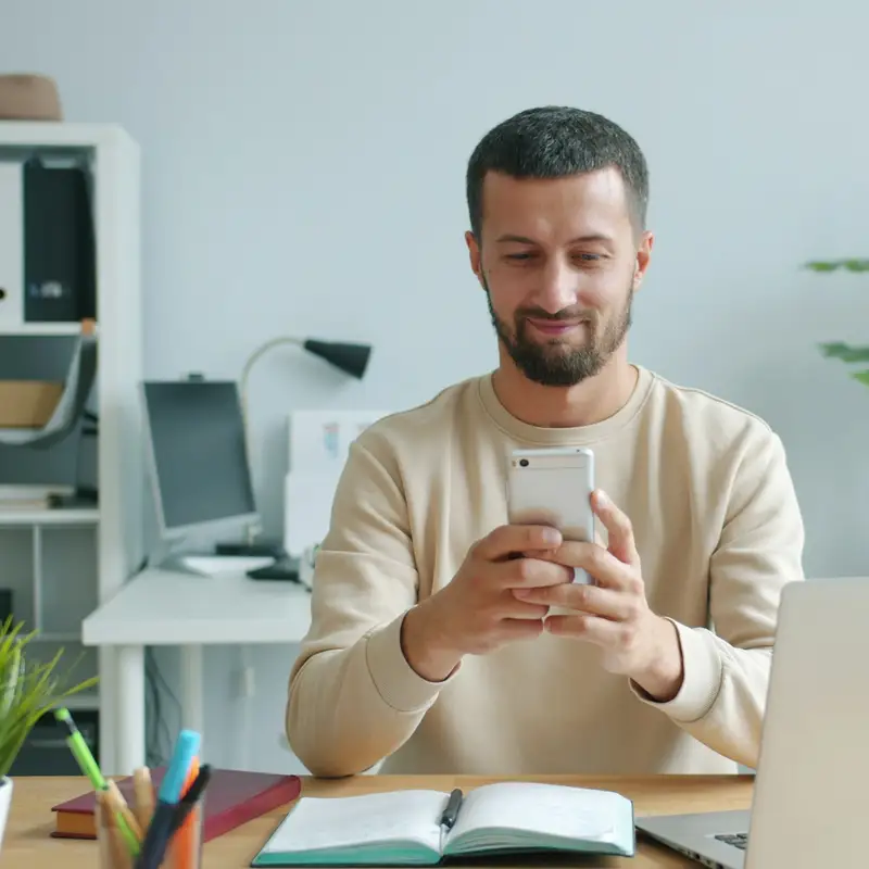 Email header photo: bearded man in a cream sweater smiling at his phone at a desk with a laptop and potted plants