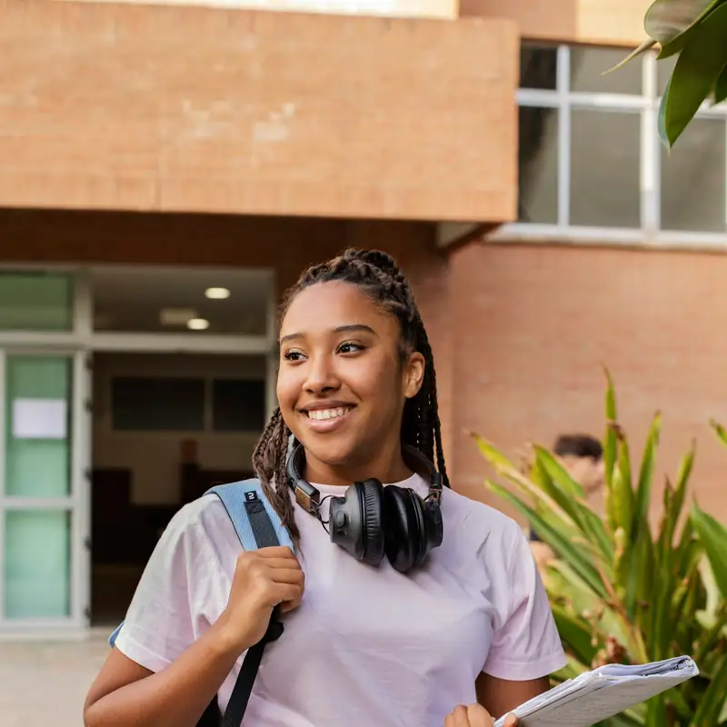 Email header photo: smiling college student with a backpack, headphones around her neck and notebooks in hand outside a brick campus building