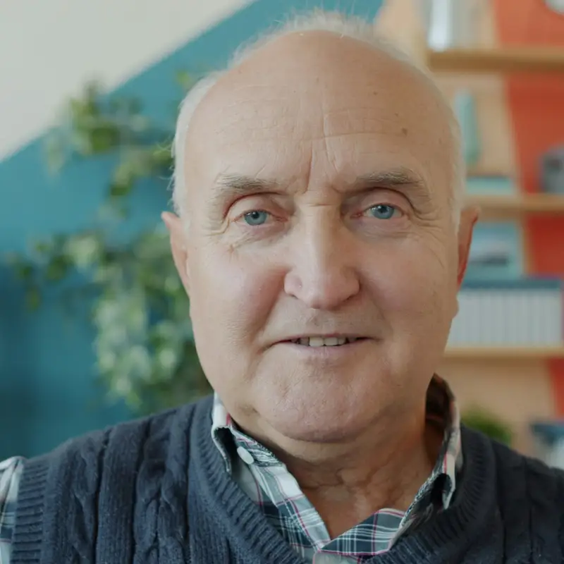Email header photo: smiling older man in a sweater vest and checked shirt sitting in a bright living room with shelves behind him