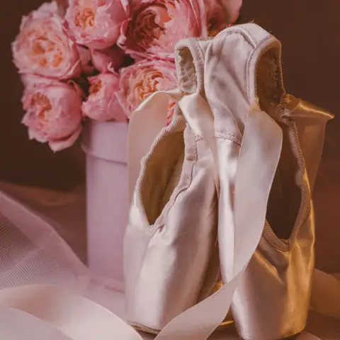 A pair of satin pointe shoes with ribbons beside a bouquet of pink peonies