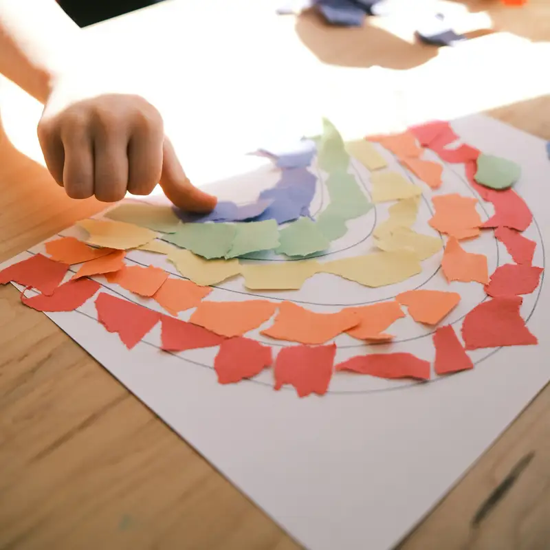 Email header photo: a child’s hand pressing colorful paper scraps onto a rainbow craft on a wooden classroom table