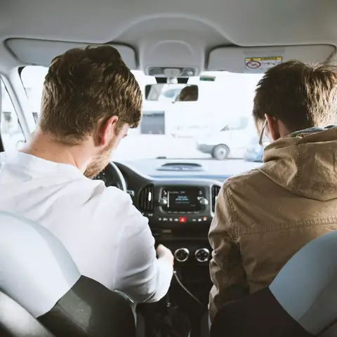 Two men in the front seats of a car seen from the back seat, one at the wheel and one in the passenger seat, on a sunny day