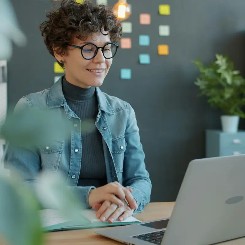 Email header photo: a smiling woman with curly hair and glasses working through a lesson on her laptop in a bright room
