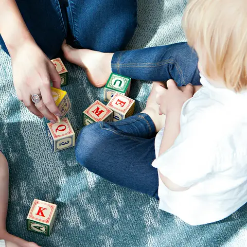 Two toddlers and a caregiver playing with wooden letter blocks on a rug