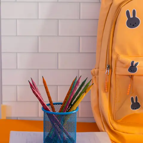 A yellow school backpack beside a stack of notebooks and a blue mesh cup of colored pencils on an orange desk