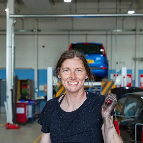 A smiling woman in a black T-shirt holding a screwdriver in an auto repair garage with a car on a lift behind her