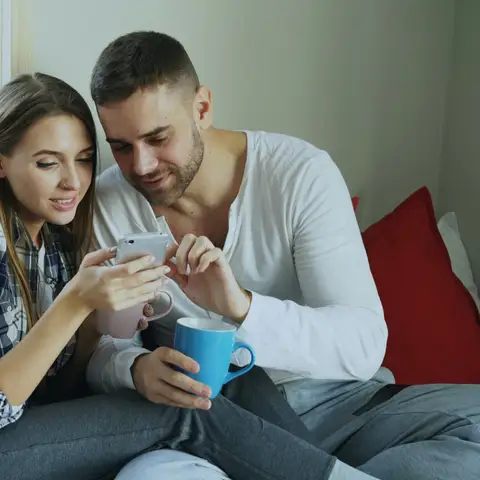 A young couple on a sofa at home looking at a phone together, one holding a coffee mug