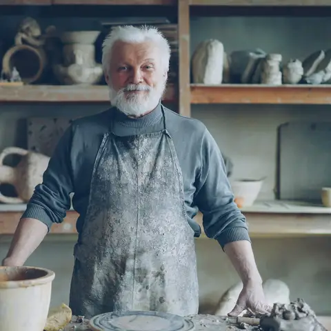A smiling gray-bearded potter in a clay-covered apron standing at his workbench in front of shelves of finished pottery