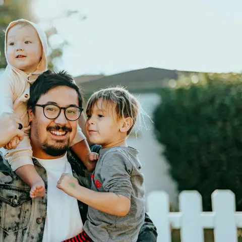 A smiling father carrying a toddler on his shoulders and a young boy in his arm, in front of a white picket fence