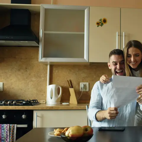 A delighted couple in their kitchen reading a letter together, the man seated at the table and the woman hugging him from behind