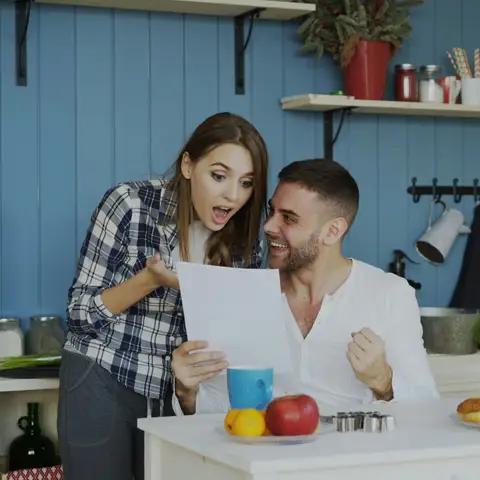 A young couple in a bright kitchen read a letter together and react with surprise and delight