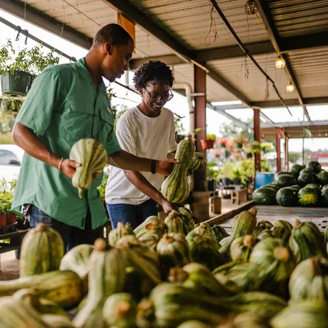 A smiling couple choosing gourds from a pile at an open-air fall market stand