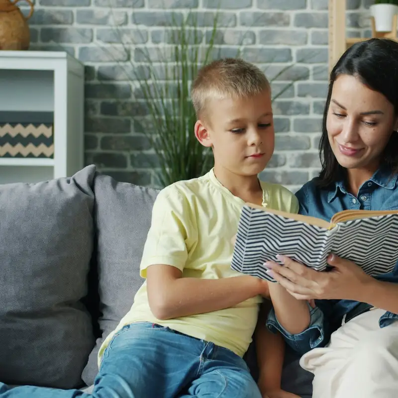 Email header photo: a smiling woman and a young boy reading a book together on a gray sofa at home