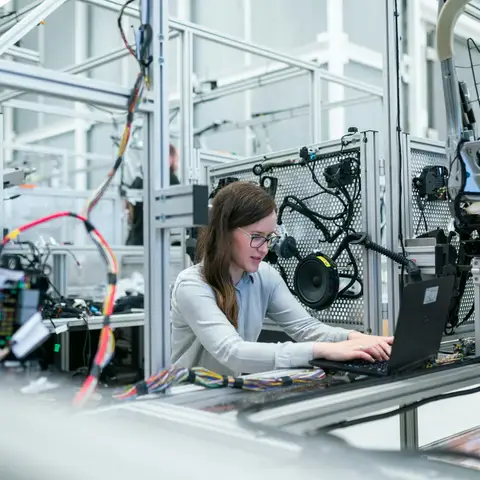 A young engineer in a light shirt working on a laptop at a test station surrounded by wiring and equipment in a bright modern plant