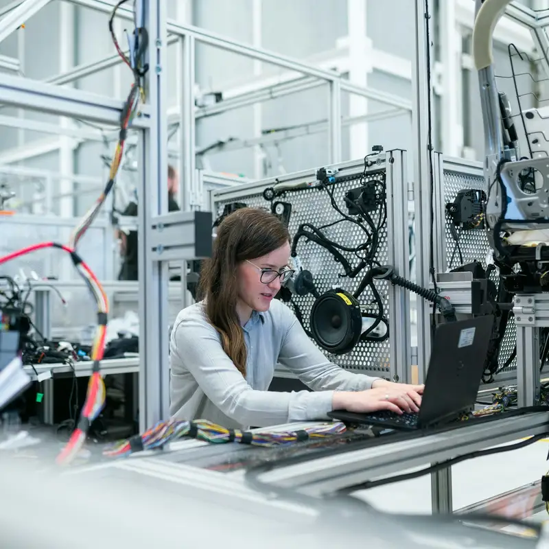 Email header photo: a young engineer in a light shirt working on a laptop at a test station surrounded by wiring and equipment in a bright modern plant