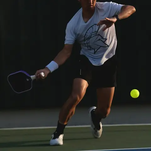 A pickleball player lunging to return a low ball on an outdoor court with a blue playing area and green surround