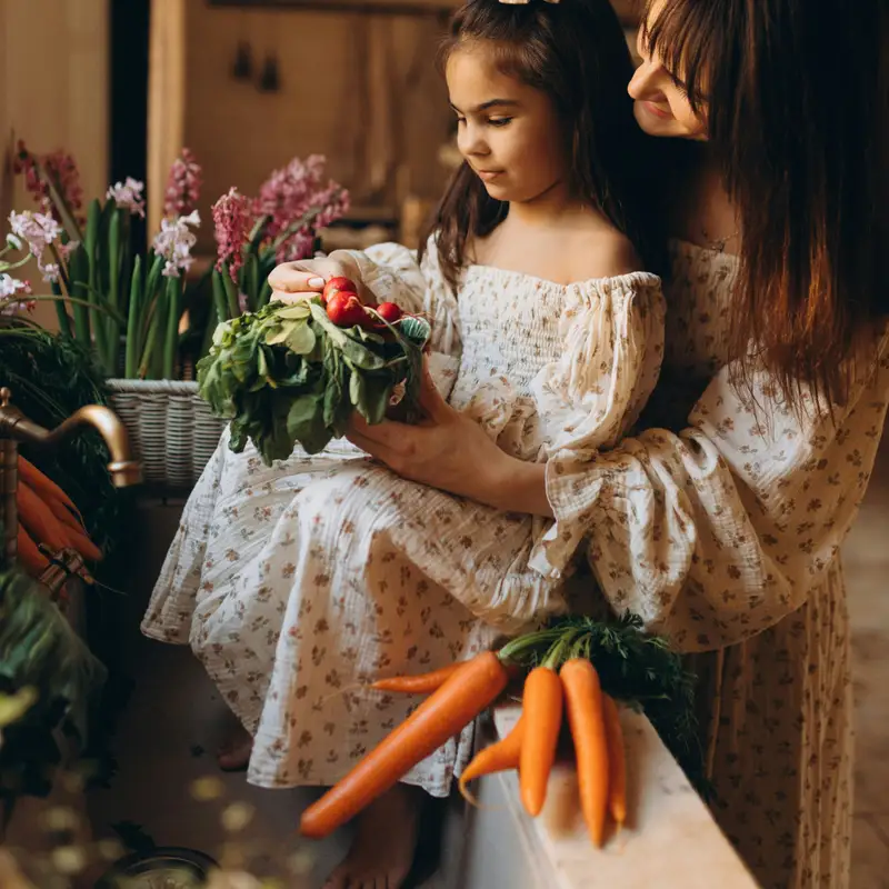 Email header photo: a mother and her young daughter in a sunny kitchen holding fresh radishes and carrots beside a vase of flowers