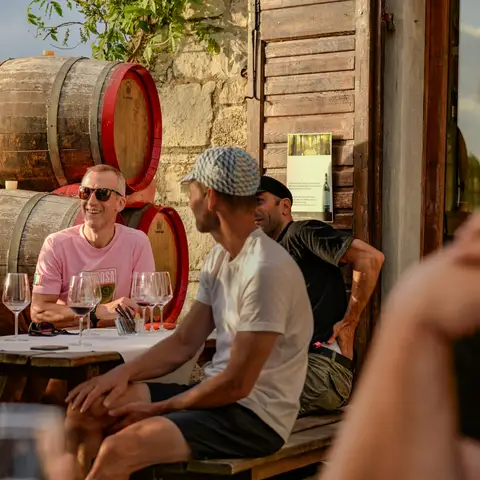 Friends laughing at a winery table with glasses of red wine, in front of stacked oak barrels in late afternoon sun
