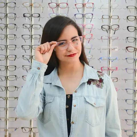 A woman in a denim jacket trying on clear frames in front of a wall of eyeglasses