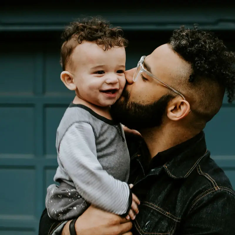 Email header photo: a father in a denim jacket kissing the cheek of his smiling toddler son in front of a gray door