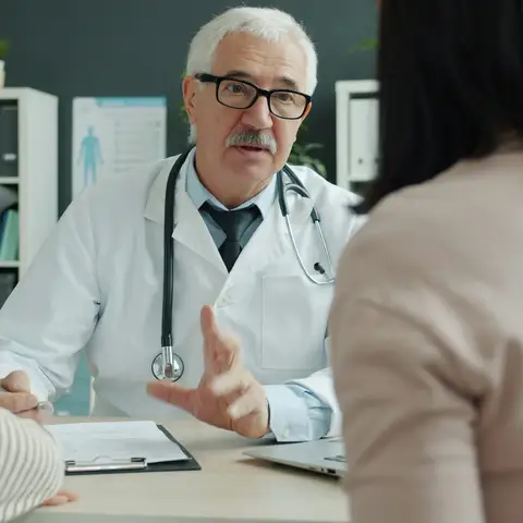 A doctor in a white coat talking with a mother and her young son across a desk in an exam office