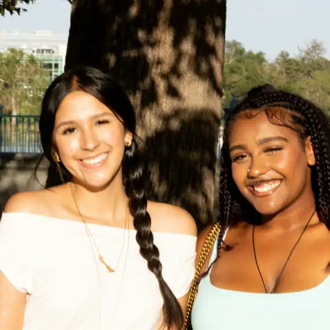 Two smiling young women standing together in front of a tree in a sunlit park